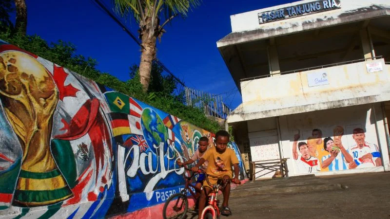 Ilustrasi warga Papua bermain sepeda berlatar belakang mural Piala Dunia 2026 di Jayapura.